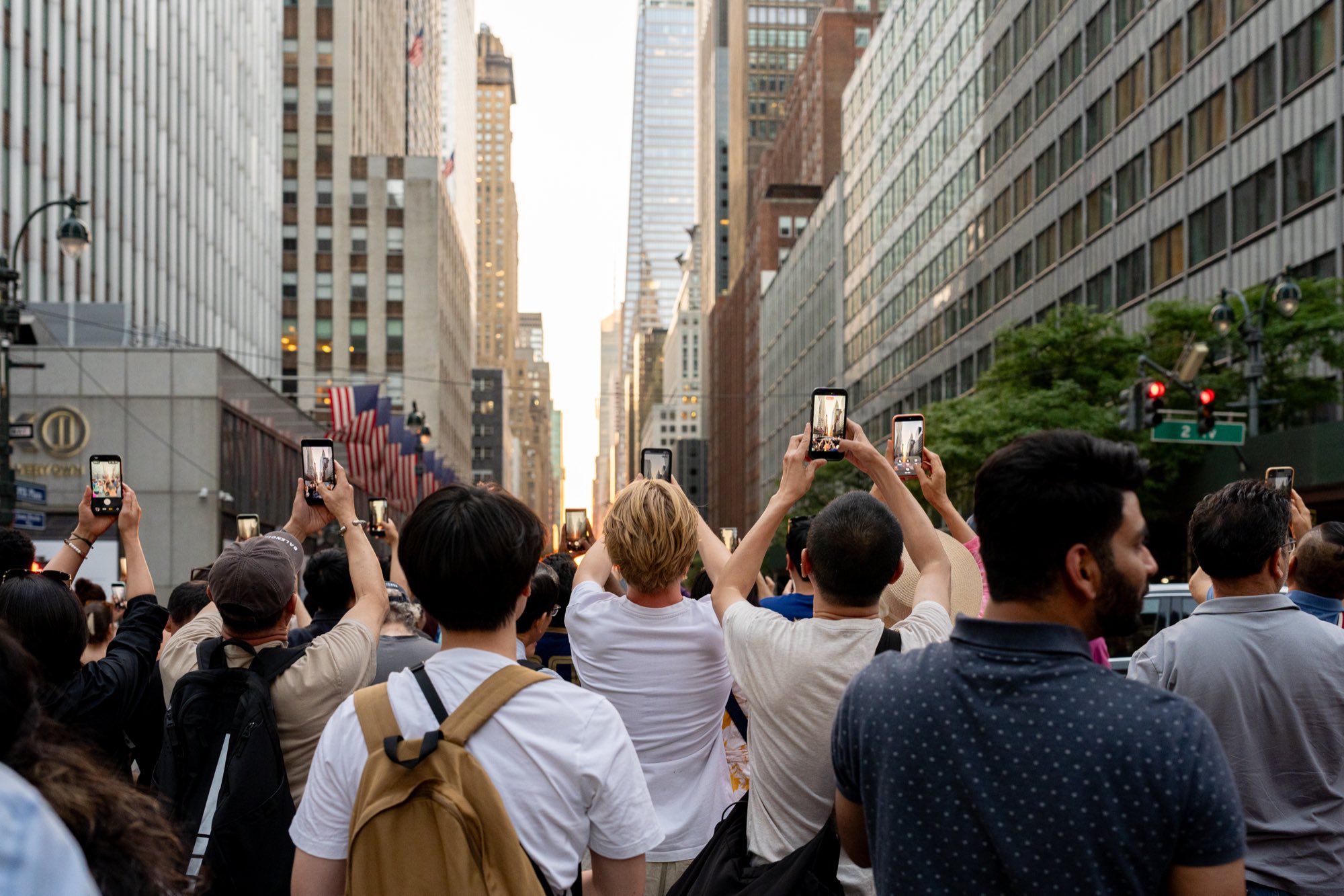 A crowd on 42nd Street holds phones overhead as the sun drops into the Manhattan grid.